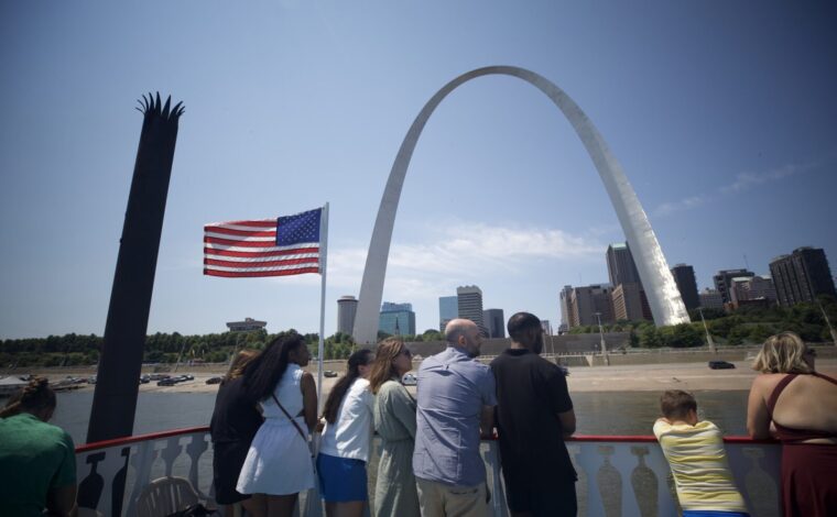 A group of people stand on the top deck of the Tom Sawyer Riverboat, looking up at the Gateway Arch during a cruise.
