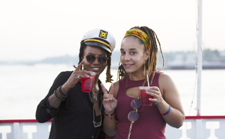 Two women holding drinks cheers and smile to the camera on the outside deck of a Groove & Spin Cruise on the Riverboats at the Gateway Arch