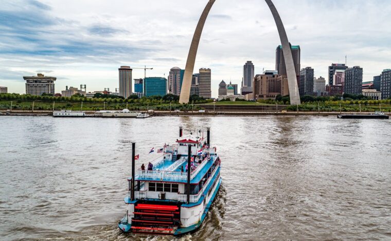 Tom Sawyer riverboat cruising on the Mississippi River with the St. Louis skyline and Gateway Arch in the background.