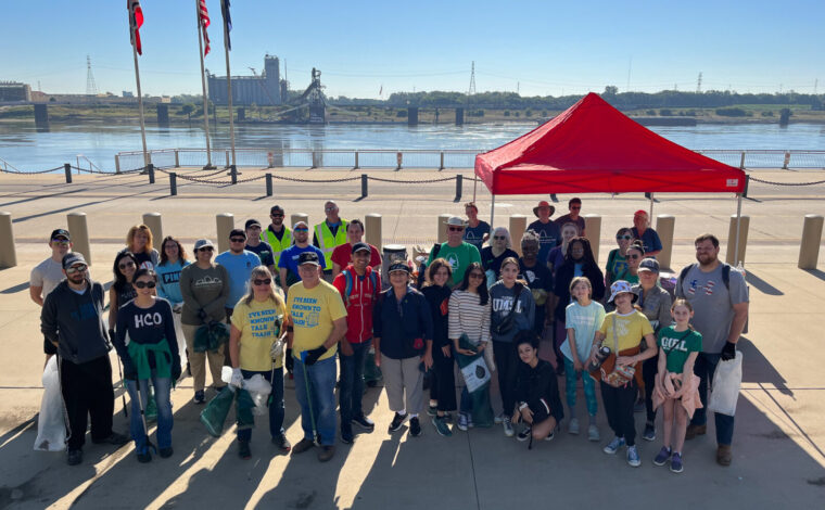 A group of about 2 dozen volunteers stand for a photo in front of the Mississippi River on the Gateway Arch National Park grounds with trash bags and pickers, ready to collect trash and clean up the St. Louis riverfront.